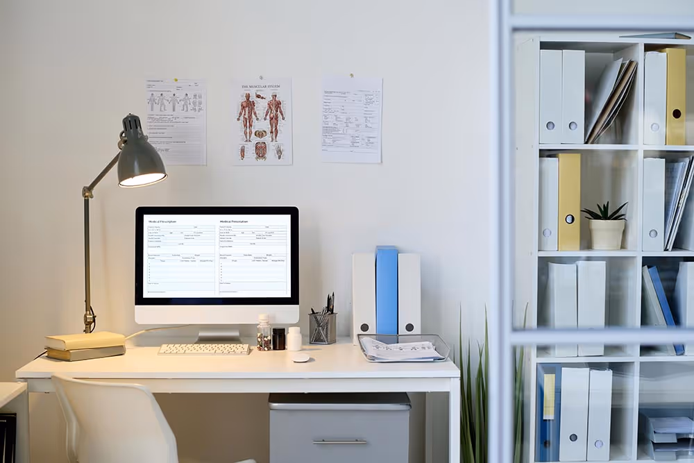 An office space with a computer turned on and a lamp turned on as well.